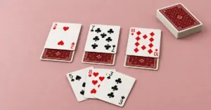 Idiot card game setup with three face-up cards over three face-down cards, a three-card hand and a draw pile on a dusty pink tabletop.