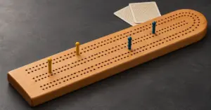 A wooden cribbage peg board with yellow and blue scoring pegs sits beside two face-down playing cards on a charcoal tabletop.