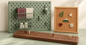 A wooden cribbage scoring board sits in front of a sage-green pegboard organising card decks and dice, beside a cork board displaying enamel pins.